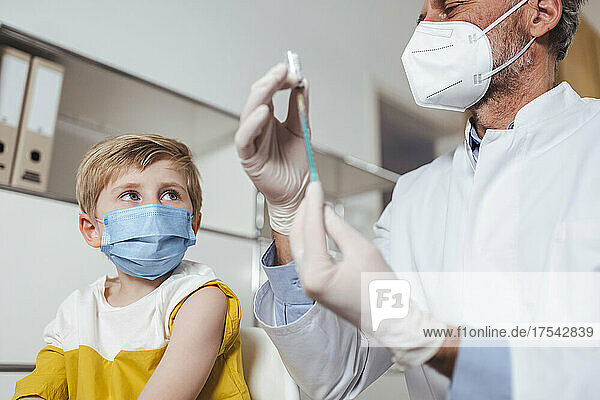 Boy wearing protective face mask looking at doctor preparing injection in center