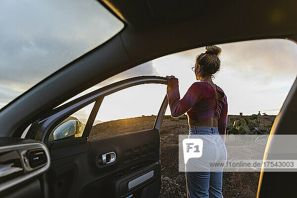 Woman standing by open car door at sunset