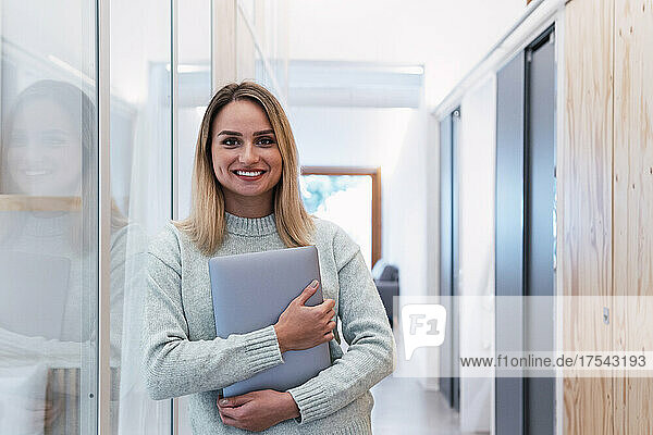 Beautiful businesswoman leaning on glass wall holding laptop in studio