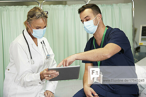 Male nurse in protective face mask explaining doctor over tablet PC in medical room