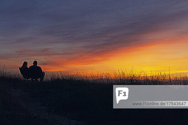 Silhouettes of man and woman sitting on bench in field at sunset