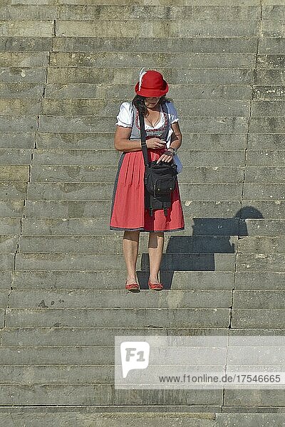 Frau im roten Dirndl steht auf Treppe und blickt ins Handy  Frau auf der Wiesn  Oktoberfest  München  Bayern  Deutschland  Europa