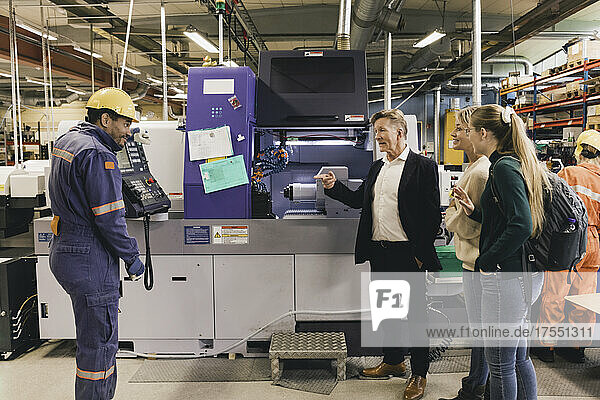 Geschäftsmann und Familie im Gespräch mit einem männlichen Arbeiter  der an einer Fertigungsmaschine in einer Fabrik steht