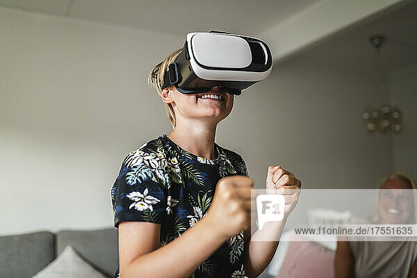 Lächelnder Junge  der ein Virtual-Reality-Headset trägt  während seine Mutter zu Hause im Hintergrund sitzt