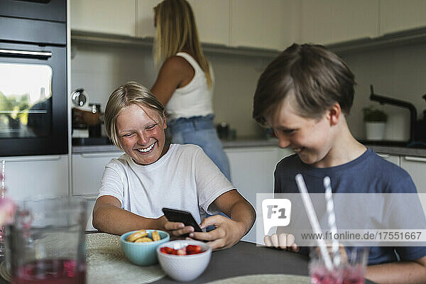 Lächelnde Brüder  die ein Smartphone benutzen  während die Mutter im Hintergrund Küchenarbeiten erledigt