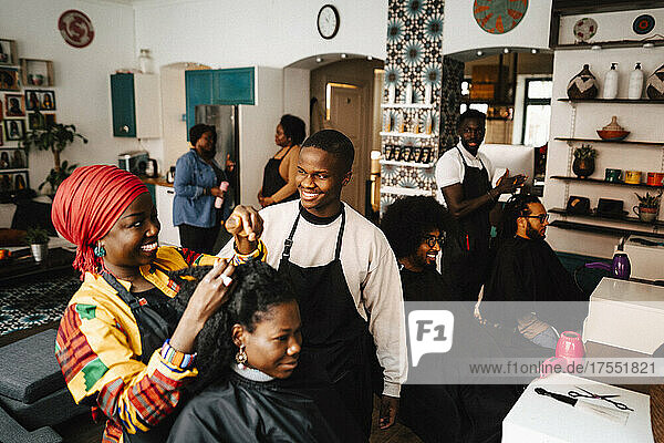 Smiling female hairdresser talking with male coworker while making locs of customer in barber shop