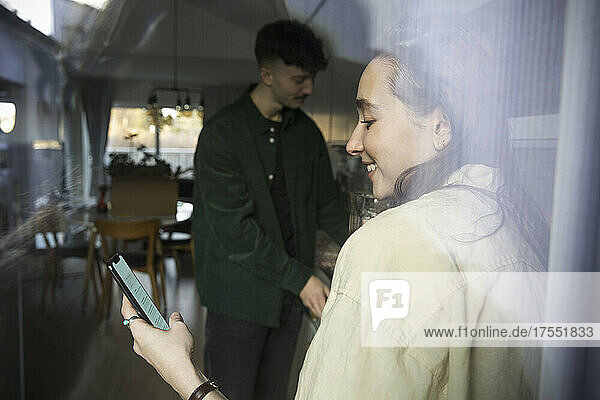 Lächelnde Frau  die ein Smartphone benutzt und neben ihrem Freund steht  gesehen durch ein Glasfenster