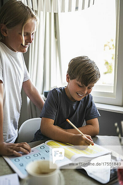 Smiling boy standing by brother doing homework at home