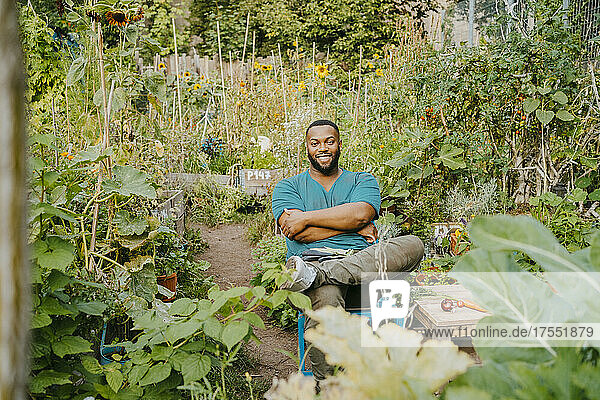 Portrait of smiling male farmer with arms crossed sitting in community garden