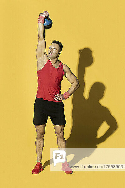 Athletischer Mann in roter Sportkleidung hebt eine Kesselglocke über Kopf vor gelbem Hintergrund