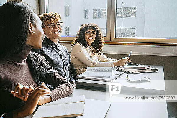 Lächelnde junge gemischtrassige Freunde sitzen zusammen am Schreibtisch in einem Universitätsklassenzimmer