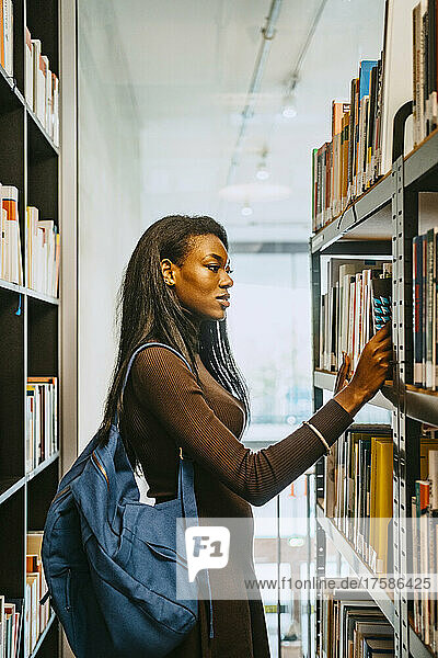 Seitenansicht einer jungen Frau mit Rucksack  die in einer Bibliothek ein Buch sucht