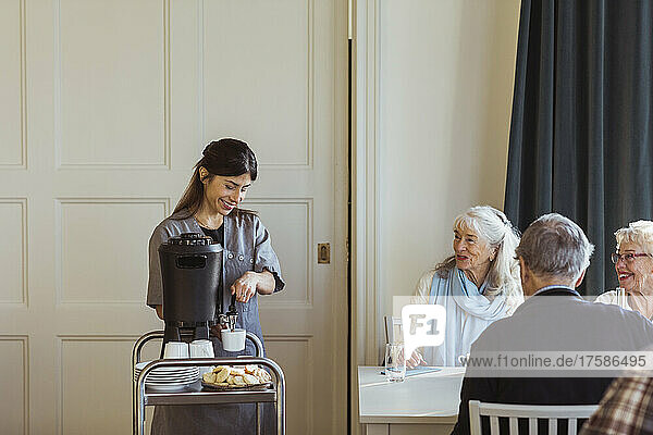 Lächelnde Mitarbeiterin im Gesundheitswesen serviert Kaffee an ältere Frauen und Männer im Altersheim