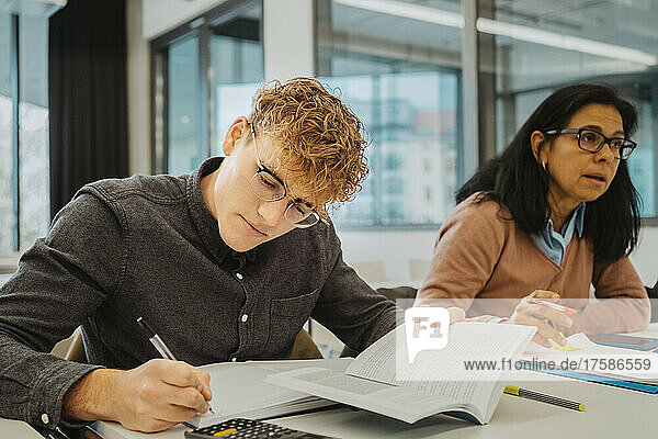 Blonder Mann schreibt  während er neben einer reifen Studentin am Schreibtisch im Klassenzimmer sitzt