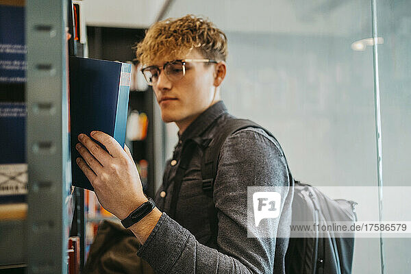Seitenansicht eines jungen Mannes mit Brille  der ein Buch in der Bibliothek einer Universität durchsucht