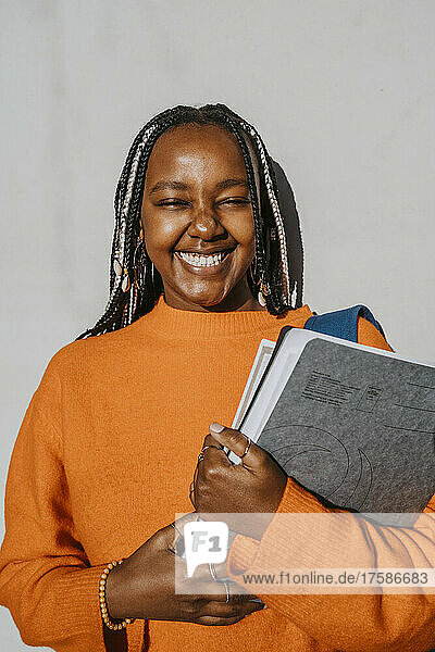 Porträt einer fröhlichen jungen Frau mit geflochtener Frisur  die ein Buch und einen Ordner vor einer grauen Wand an einem sonnigen Tag hält