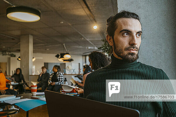 Thoughtful young bearded man sitting with laptop in university cafeteria