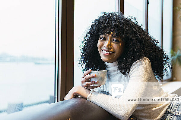 Smiling curly haired woman with coffee cup leaning on sofa at cafe