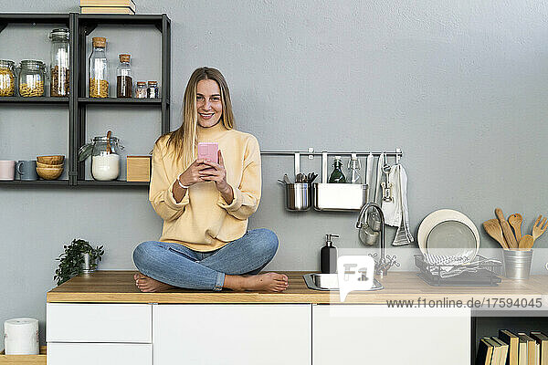 Smiling woman with mobile phone sitting on kitchen counter at home