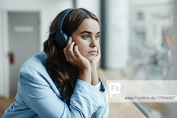 Thoughtful businesswoman with head in hand listening music through wireless headphones in office