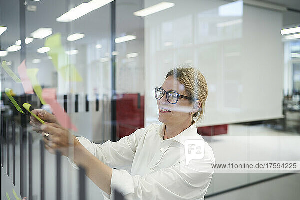 Smiling businesswoman sticking adhesive note on glass wall in office