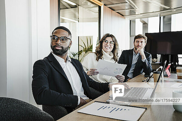 Young businessman wearing eyeglasses sitting by colleagues at conference table in board room