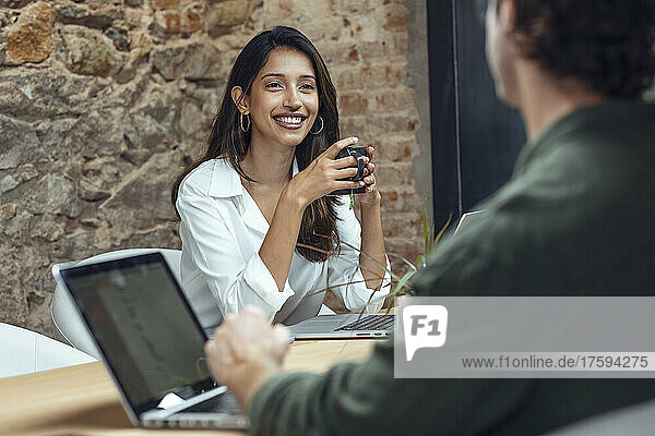 Lächelnde Geschäftsfrau hält Kaffeetasse in der Hand und diskutiert mit Geschäftsmann am Schreibtisch im Büro