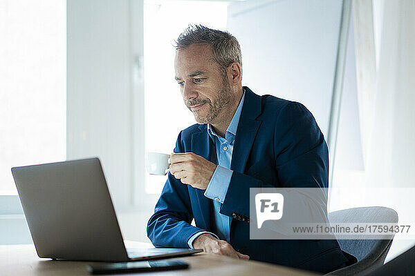 Businessman holding coffee cup using laptop at desk in office
