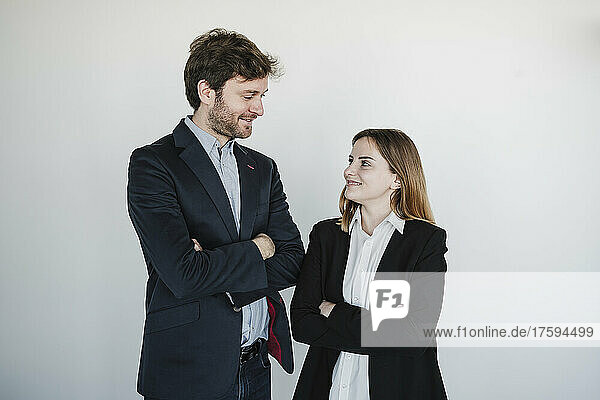 Smiling businesswoman and businessman standing with arms crossed looking at each other in front of wall