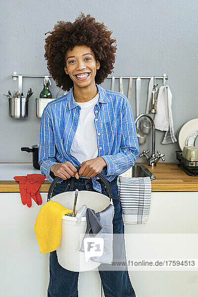 Smiling young Afro woman holding bucket with cleaning equipment in kitchen