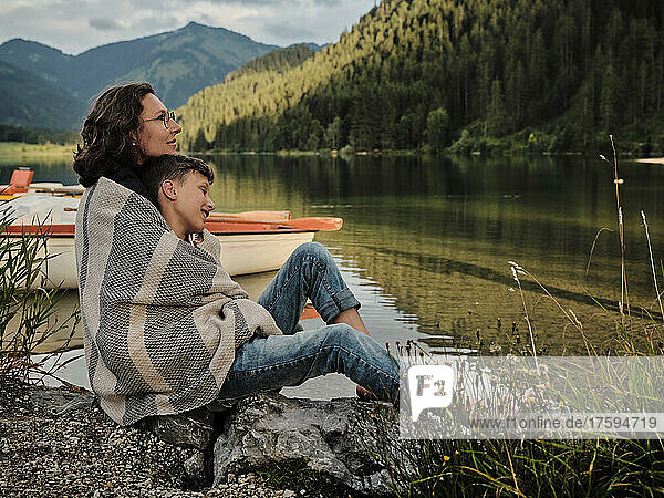 Mother and son wrapped in blanket sitting at lakeshore