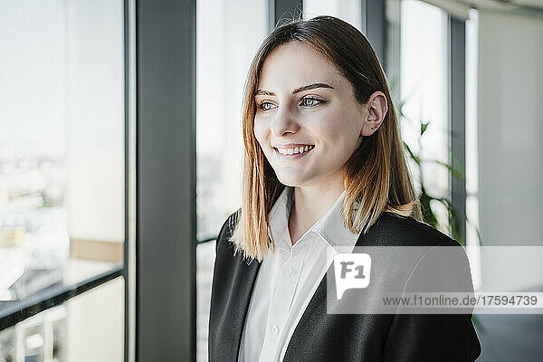 Smiling young beautiful businesswoman standing by window in office
