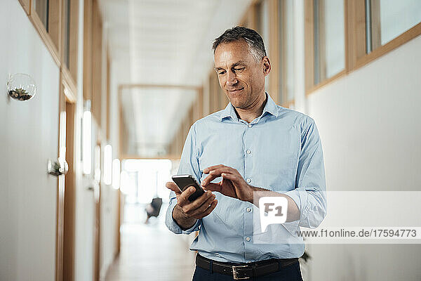 Mature businessman using smart phone at corridor in office
