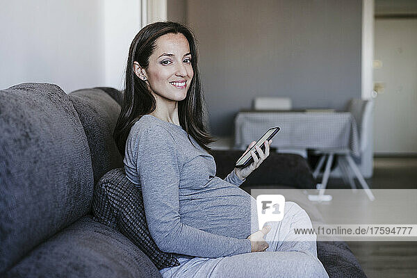 Lächelnde schwangere Frau mit Smartphone sitzt zu Hause auf dem Sofa