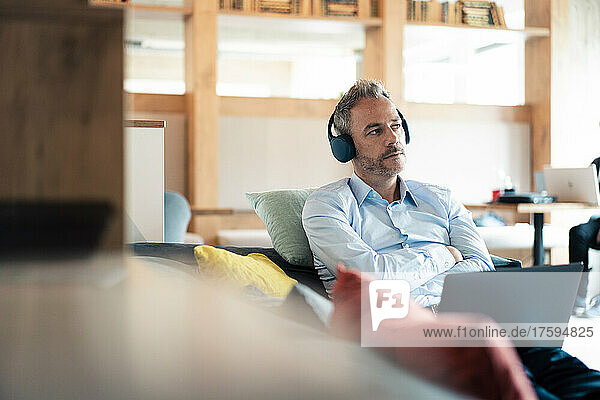 Thoughtful businessman listening music sitting with arms crossed at office