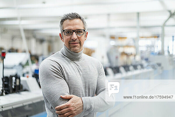 Smiling businessman wearing eyeglasses standing with arms crossed in factory