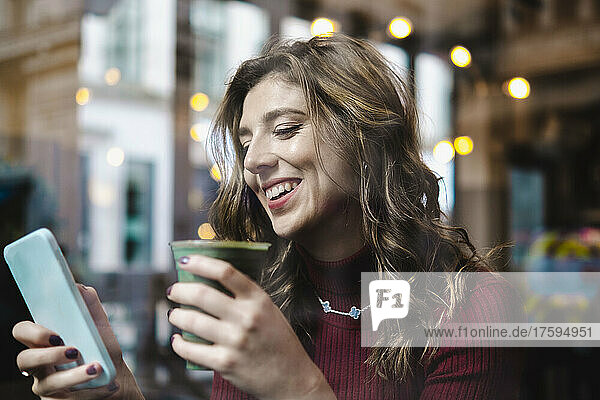 Lächelnde Frau mit Einwegkaffeetasse und Smartphone im Café