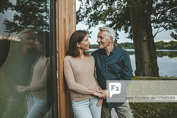 Smiling couple holding hands looking at each other by glass wall in garden