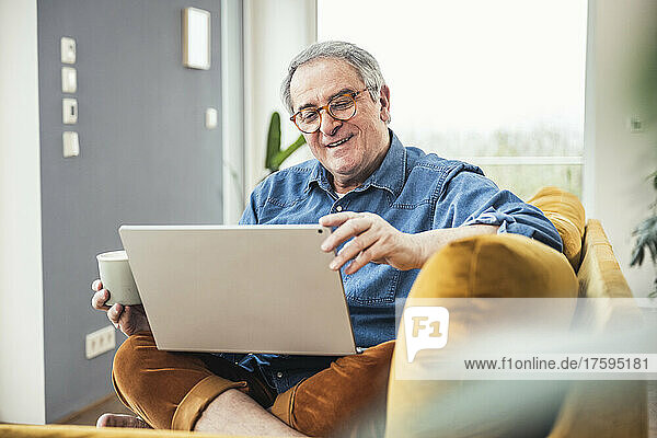 Senior man holding coffee cup watching video on laptop in living room at home