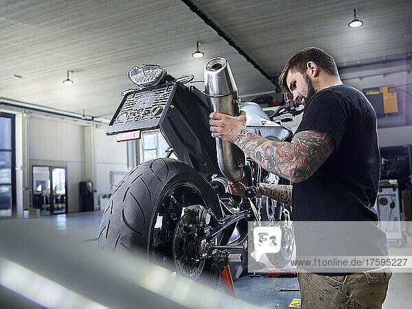Young tattooed mechanic examining exhaust pipe of motorcycle in workshop