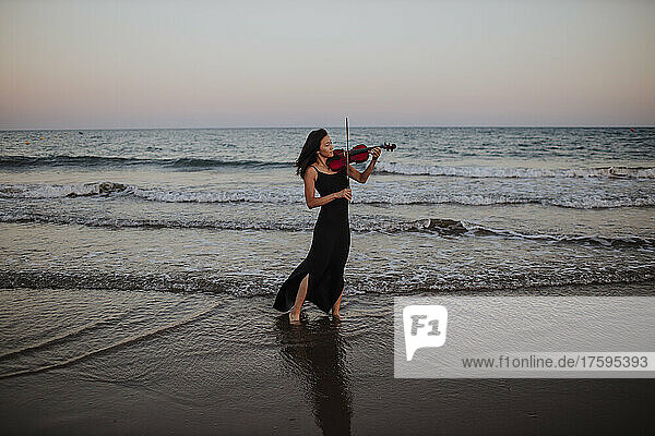 Frau spielt Geige und steht bei Sonnenuntergang vor dem Meer