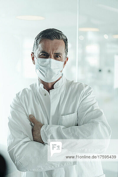 Scientist with protective face mask leaning on glass wall at medical clinic