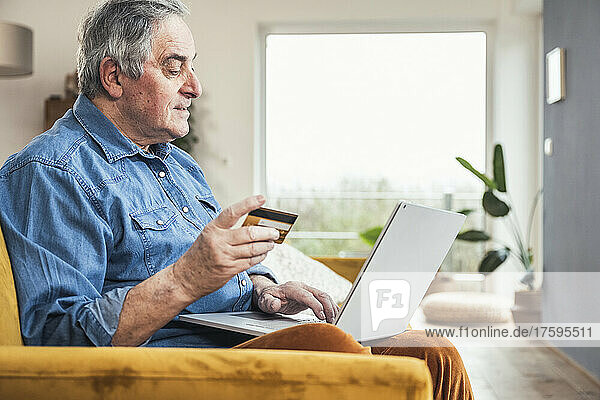 Senior man with credit card making online payment through laptop in living room