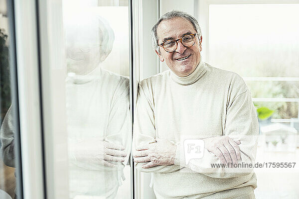 Smiling senior man with arms crossed standing by window at home