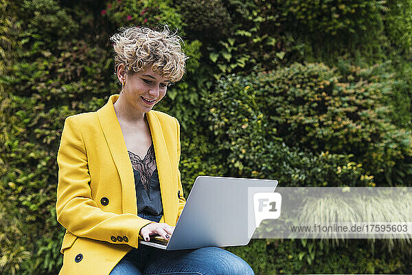 Smiling businesswoman in yellow jacket working on laptop at park