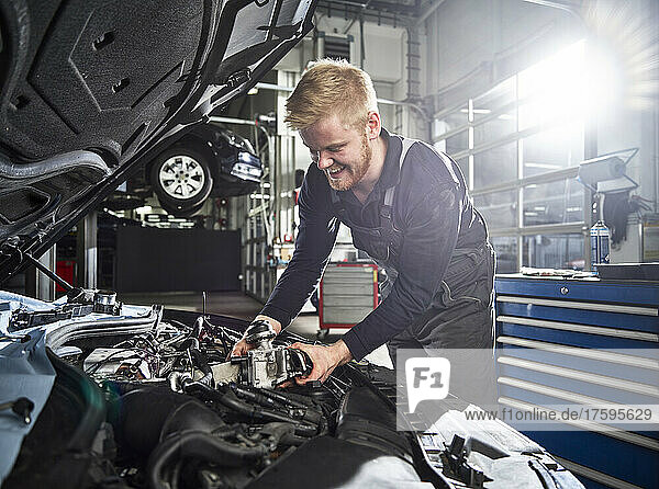 Smiling young blond mechanic repairing car engine in auto shop