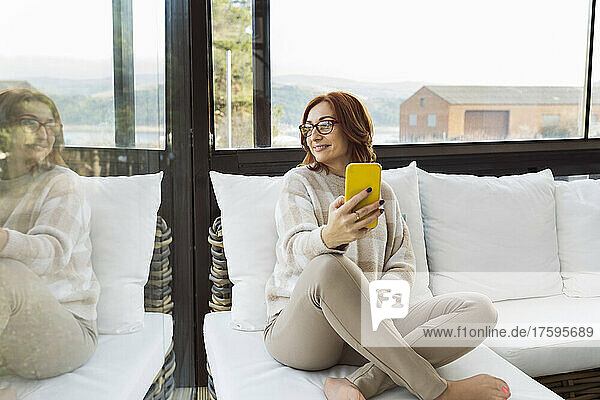 Smiling woman with smart phone looking though window at home
