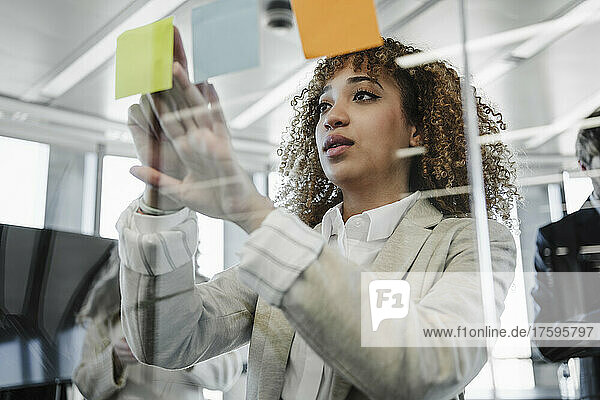 Junge Geschäftsfrau mit braunem lockigem Haar klebt im Büro Klebezettel an die Glaswand