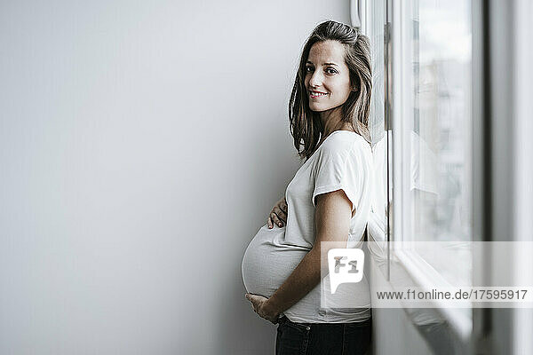 Lächelnde schwangere Frau lehnt zu Hause am Fenster