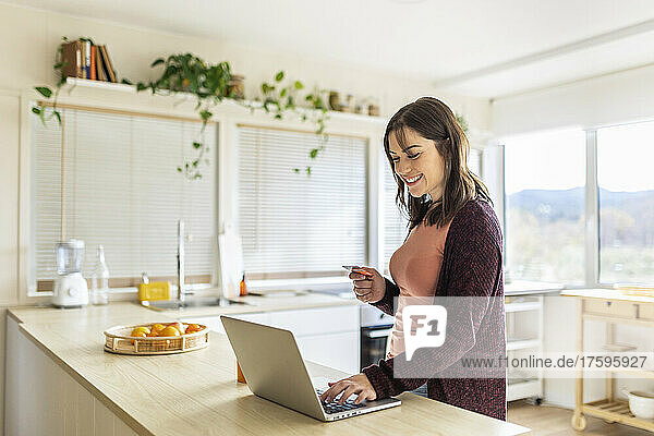 Businesswoman doing online shopping on laptop holding credit card in kitchen at home